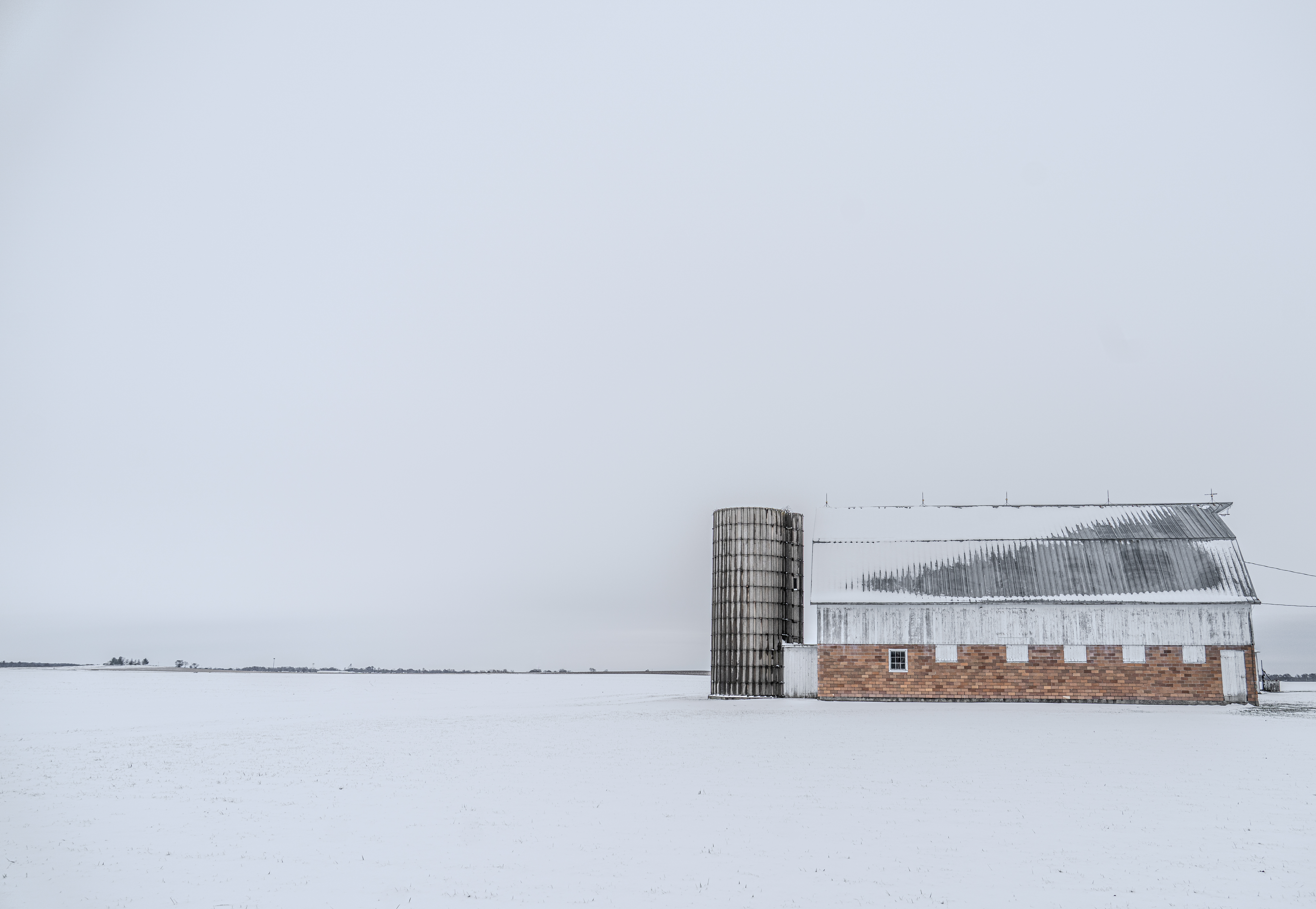 A barn and silo stand against a gray sky and snowy background